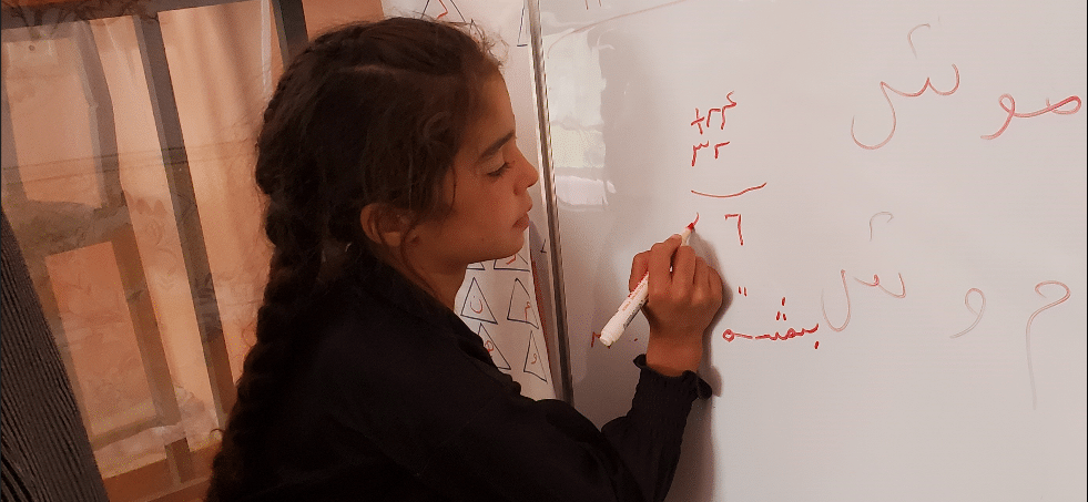 A young girl with dark hair in a braid writes a mathematical equation on a whiteboard.