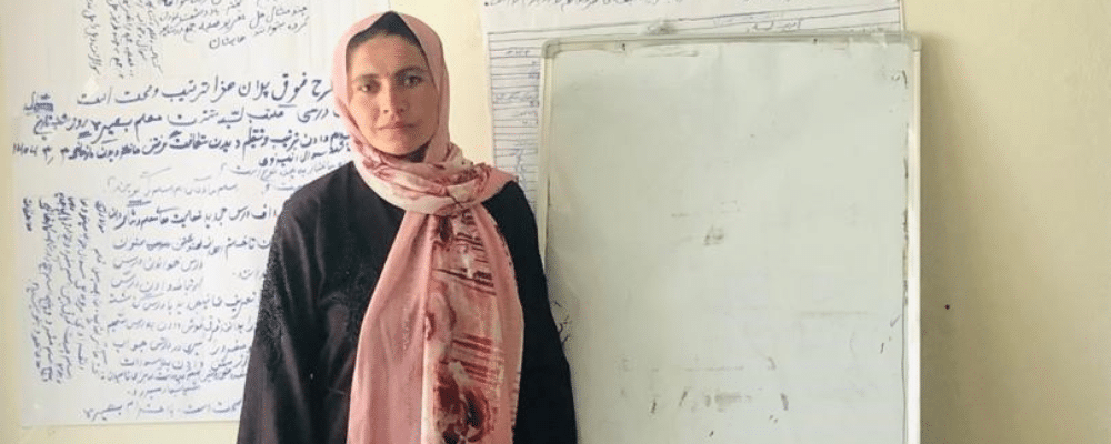 A woman wearing a pink headscarf stands in front of her classroom looking at the camera.
