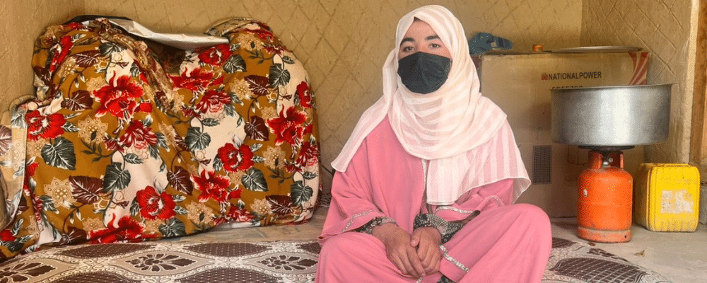 A woman wearing a beige hijab and pink robe sits before the camera.