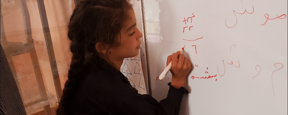 A young girl with dark hair in a braid writes a mathematical equation on a whiteboard.